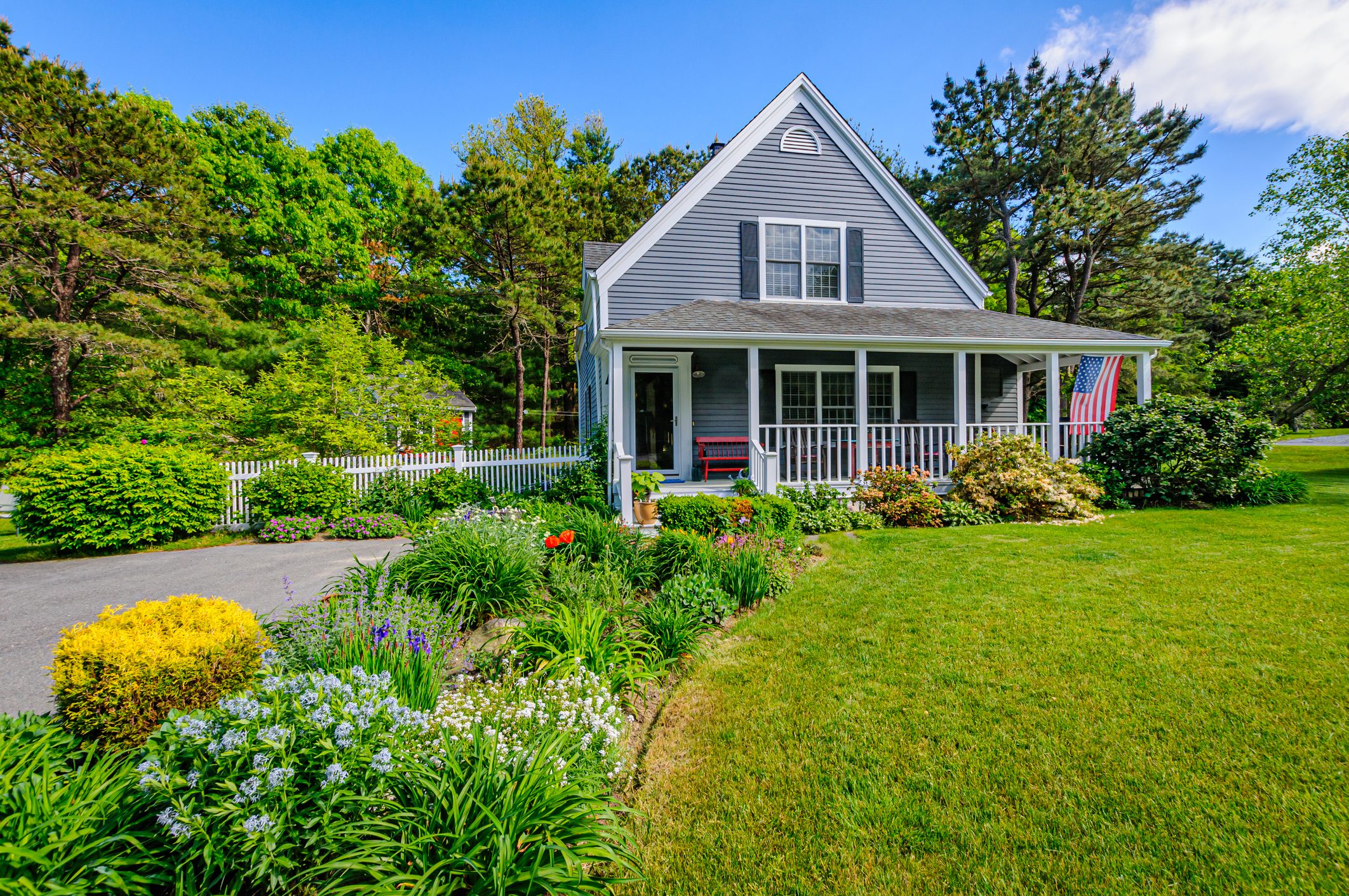 Springtime gardens along the driveway of a small single family home on Cape Cod with a flag flying from the front porch,  and the surrounding grounds,  on a late May afternoon with copy space in a clear sky area.